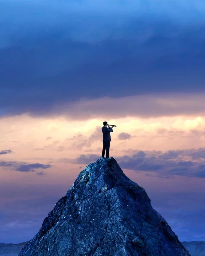 A person in a suit stands on top of a rocky mountain peak, looking through a telescope at a dramatic, colorful sky with clouds at sunset or sunrise. - Lead Experts