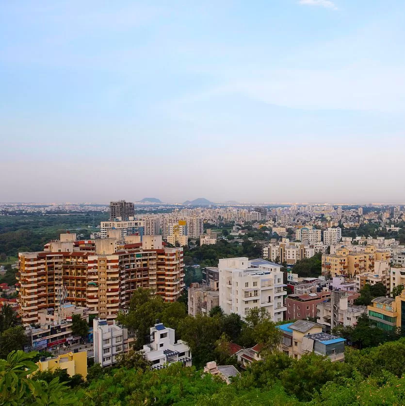 A cityscape view shows numerous mid-rise buildings surrounded by greenery, with a sprawling urban area extending into the distance under a partly cloudy sky. Distant hills are visible on the horizon. - Lead Experts