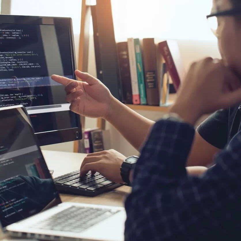 Two people sit at a desk with a laptop and desktop computer, discussing code displayed on both screens. One person points at the monitor while the other observes. Bookshelves are visible in the background. - Lead Experts