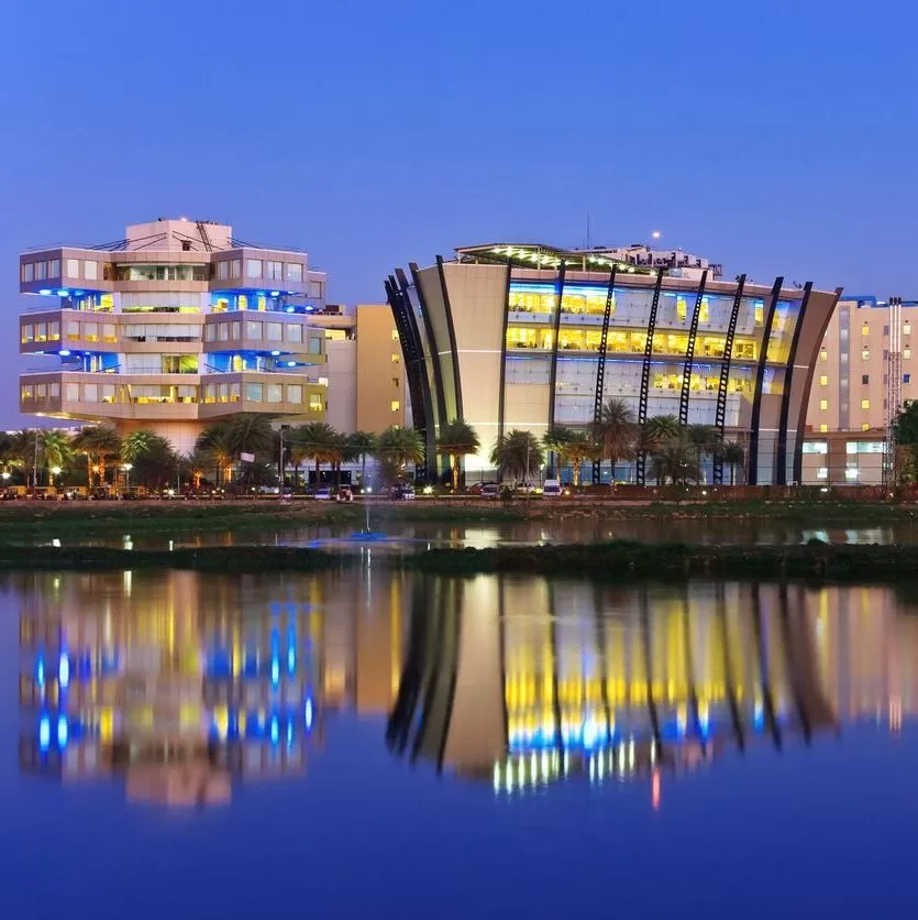 Modern office buildings with illuminated windows are reflected in a calm lake at dusk, with palm trees lining the waterfront and a clear blue sky in the background. - Lead Experts