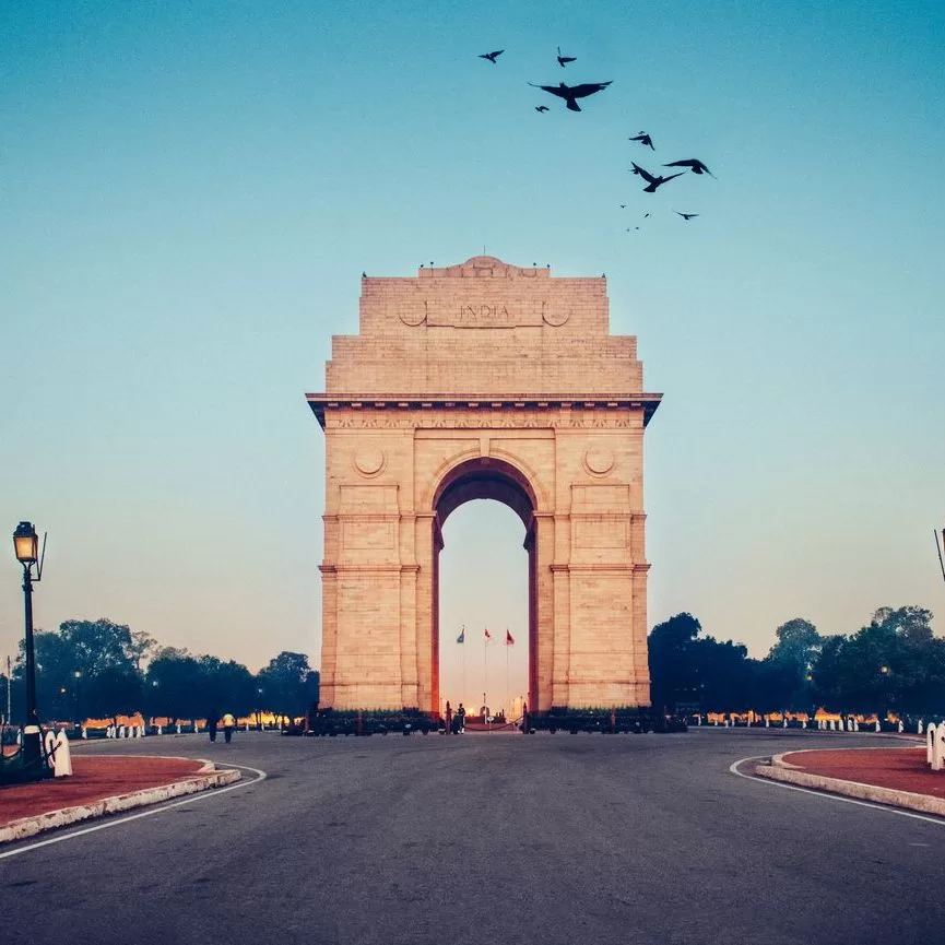 India Gate, a large stone war memorial arch, stands at the center of a wide road at sunrise or sunset, with birds flying above and trees silhouetted in the background under a clear blue sky. - Lead Experts
