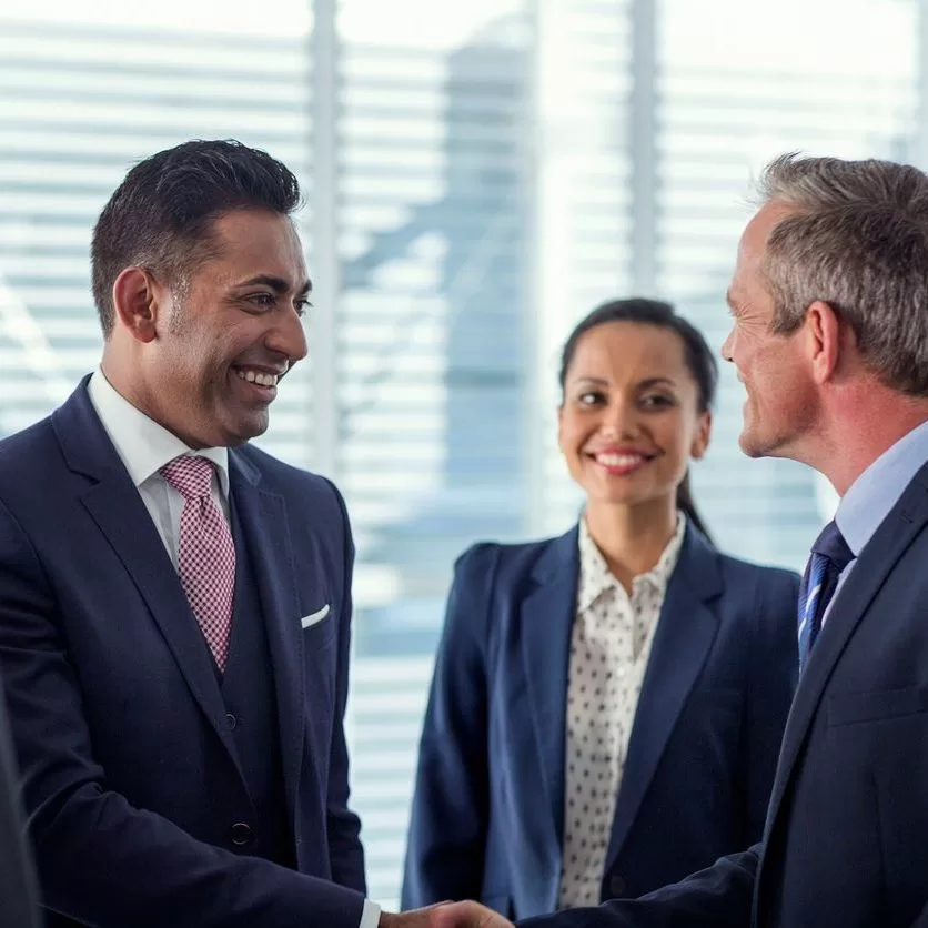 Two businessmen in suits shake hands and smile, while a businesswoman in a suit stands in the background smiling. The setting appears to be a modern office with large windows and blinds. - Lead Experts