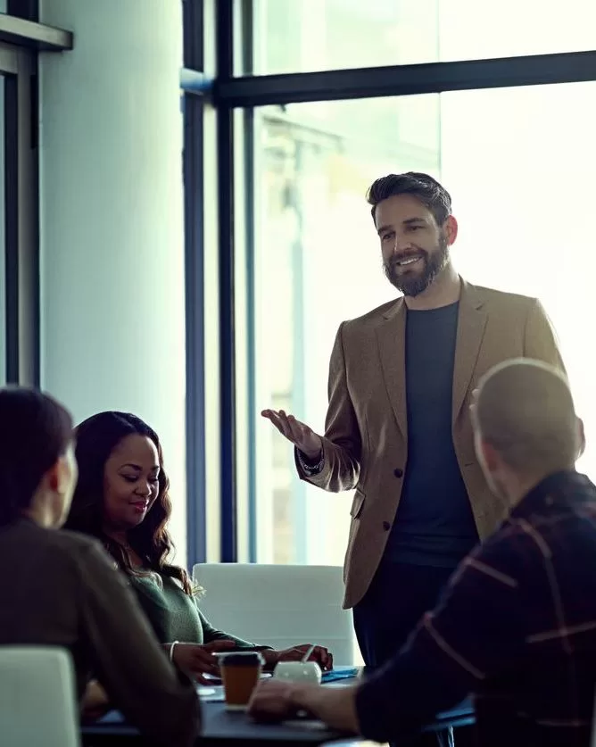 A man in a brown blazer is standing and speaking to three seated colleagues in a bright, modern office with large windows, introducing lead experts during a casual meeting or discussion. - Lead Experts