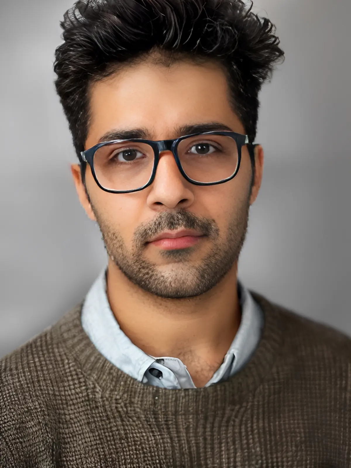 A young man with short dark hair, wearing black glasses, a light blue collared shirt, and a brown sweater, looks seriously at the camera against a plain gray background. - Lead Experts