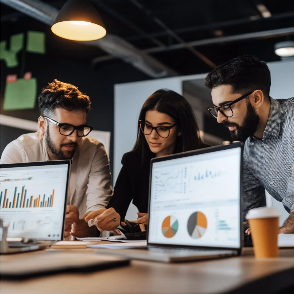 Three people collaborate at an office desk, reviewing documents and data on paper. Two laptops in the foreground display hire SEO charts and graphs. The workspace is modern, with warm lighting and post-it notes on the wall. - Lead Experts