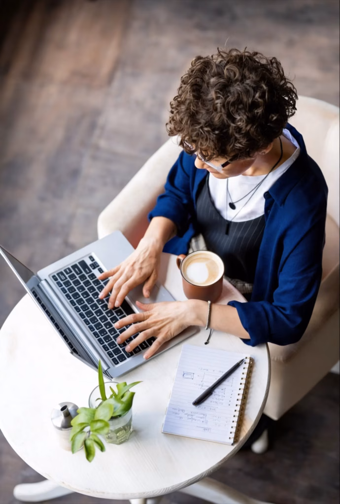 A person with curly hair sits at a round table, typing on a laptop—perhaps working on content writing services. A cup of coffee, notepad with pen, small plant, and salt shaker are also on the table. The scene is viewed from above. - Lead Experts